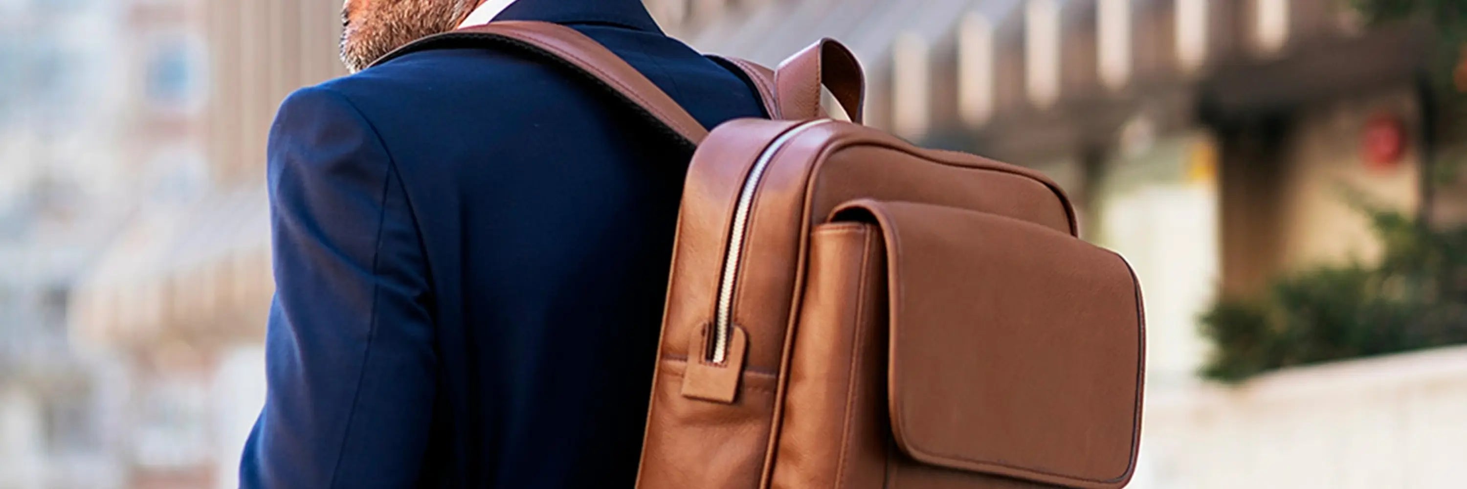Homme élégant en costume bleu portant un sac à dos homme en cuir marron, marchant en ville dans un environnement urbain chic, illustrant un équilibre entre style professionnel et praticité.